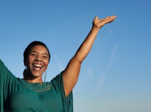 Head and upper body of a person with dark skin and dark hair, smiling, one arm raised, set against blue sky.